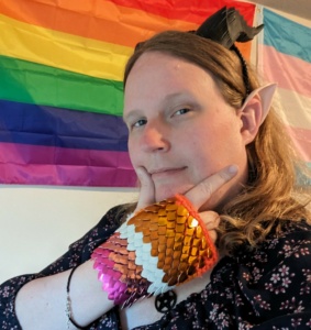 Elizabeth wearing Dragon horns, elf ears, lesbian pride dragon gloves. Standing in front of a classic LGBT pride flag and Trans pride flag.